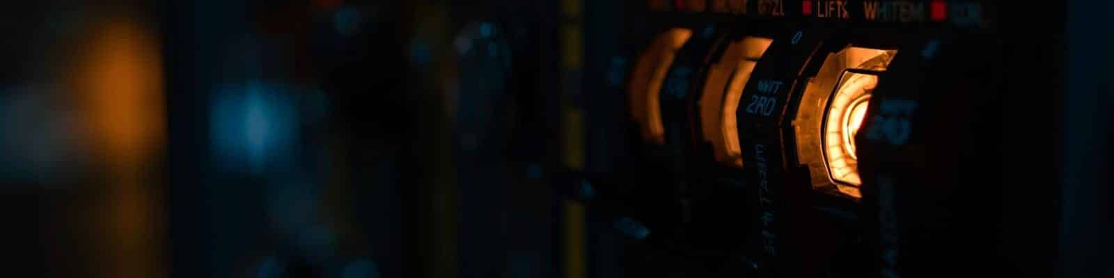 Close de um painel elétrico escuro com interruptores acesos em laranja, destacando detalhes técnicos iluminados.