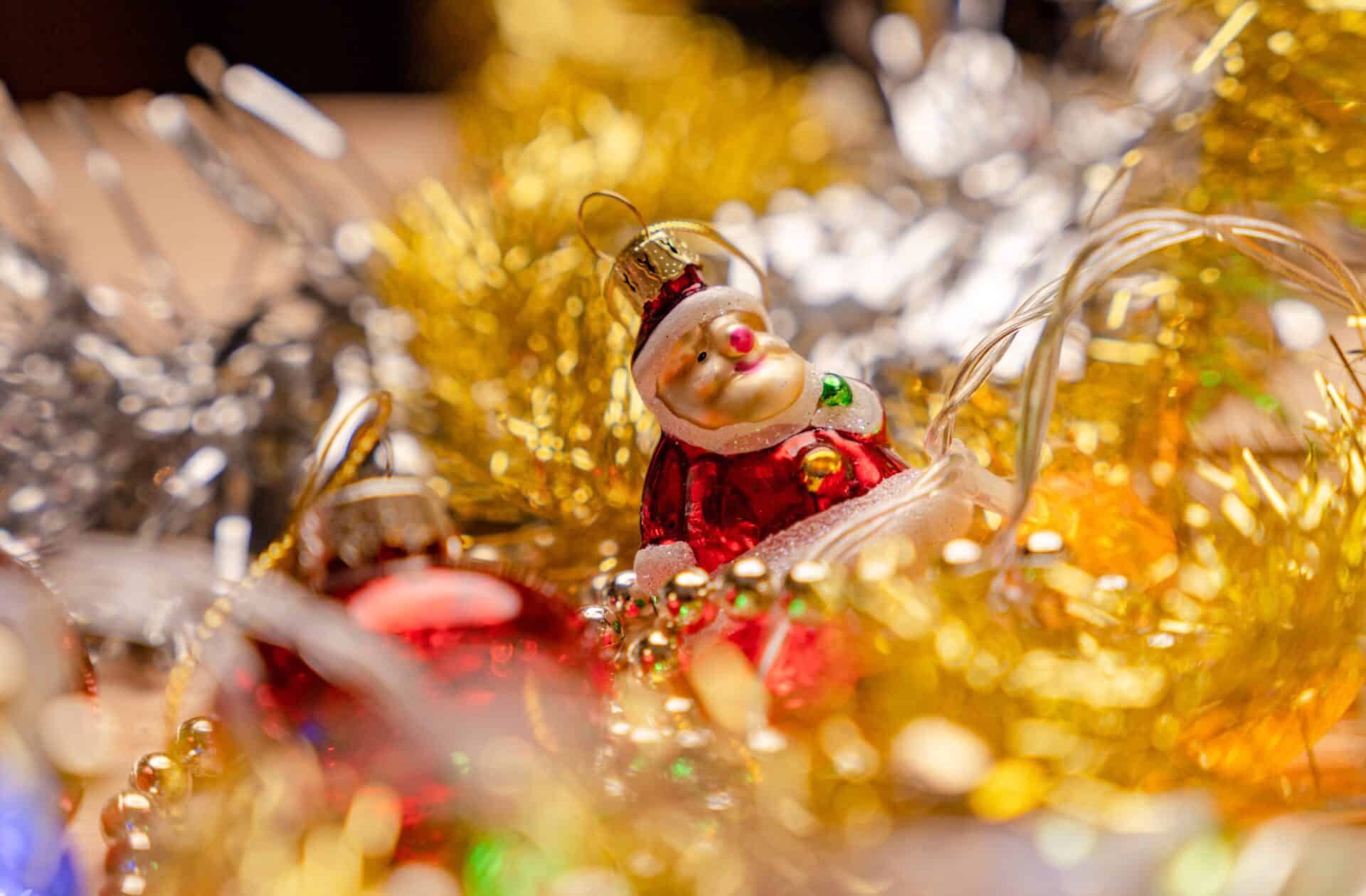 fotografia em close-up de enfeites de Natal com foco seletivo. No centro, destaca-se uma pequena figura de Papai Noel vermelha e dourada, cercada por festões brilhantes em tons de ouro e prata.