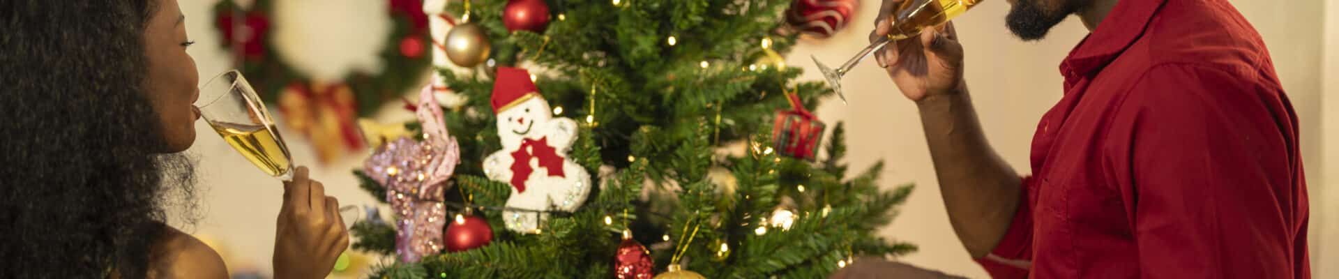 Um casal de jovens negros celebra o Natal em uma sala decorada. À esquerda, uma mulher de perfil segura uma taça de espumante, enquanto à direita, um homem vestindo uma camisa vermelha bebe de sua taça