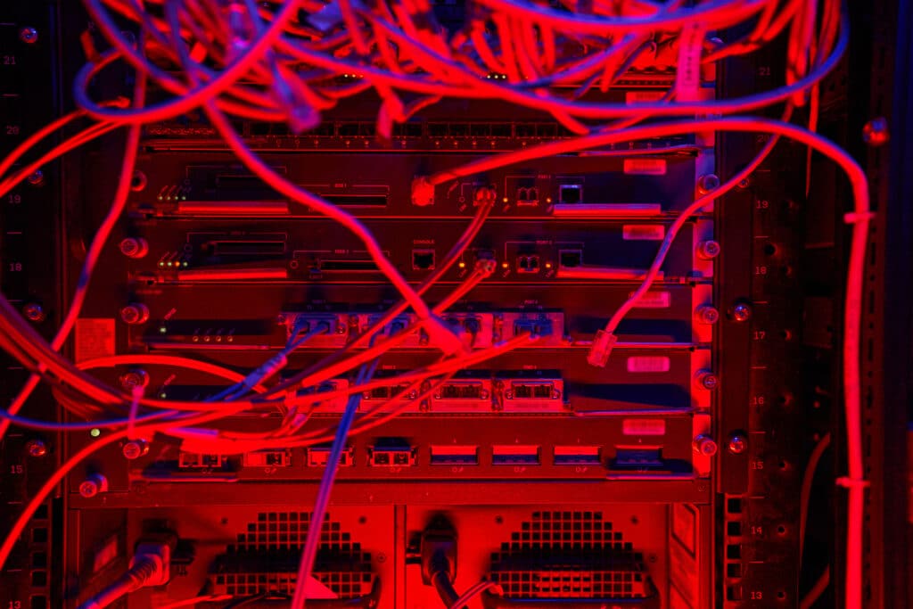 Close de um rack de servidores em um data center, iluminado por uma luz vermelha intensa. Vários cabos de rede e fibras ópticas emaranhados conectam-se às portas dos equipamentos, criando uma composição visual complexa de fios e luzes indicadoras.