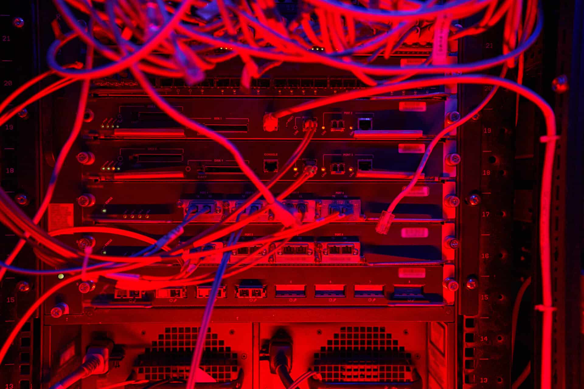 Close de um rack de servidores em um data center, iluminado por uma luz vermelha intensa. Vários cabos de rede e fibras ópticas emaranhados conectam-se às portas dos equipamentos, criando uma composição visual complexa de fios e luzes indicadoras.