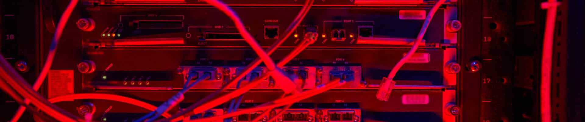 Close de um rack de servidores em um data center, iluminado por uma luz vermelha intensa. Vários cabos de rede e fibras ópticas emaranhados conectam-se às portas dos equipamentos, criando uma composição visual complexa de fios e luzes indicadoras.