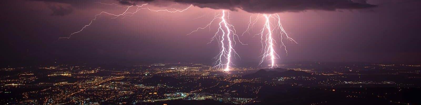 Tempestade noturna com múltiplas descargas elétricas iluminando cidade ao fundo, evidenciando riscos para redes elétricas urbanas.