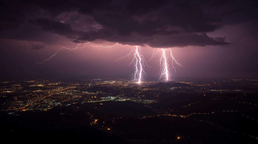 Tempestade noturna com múltiplas descargas elétricas iluminando cidade ao fundo, evidenciando riscos para redes elétricas urbanas.