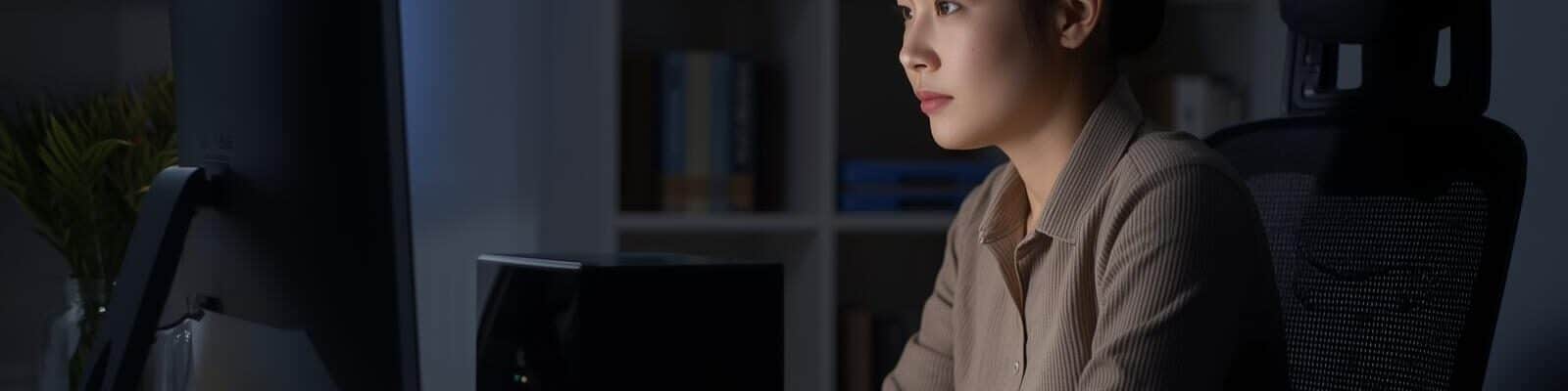 Mulher concentrada trabalhando em um computador desktop em um escritório doméstico escuro, com um nobreak preto brilhante visível ao lado do monitor.
