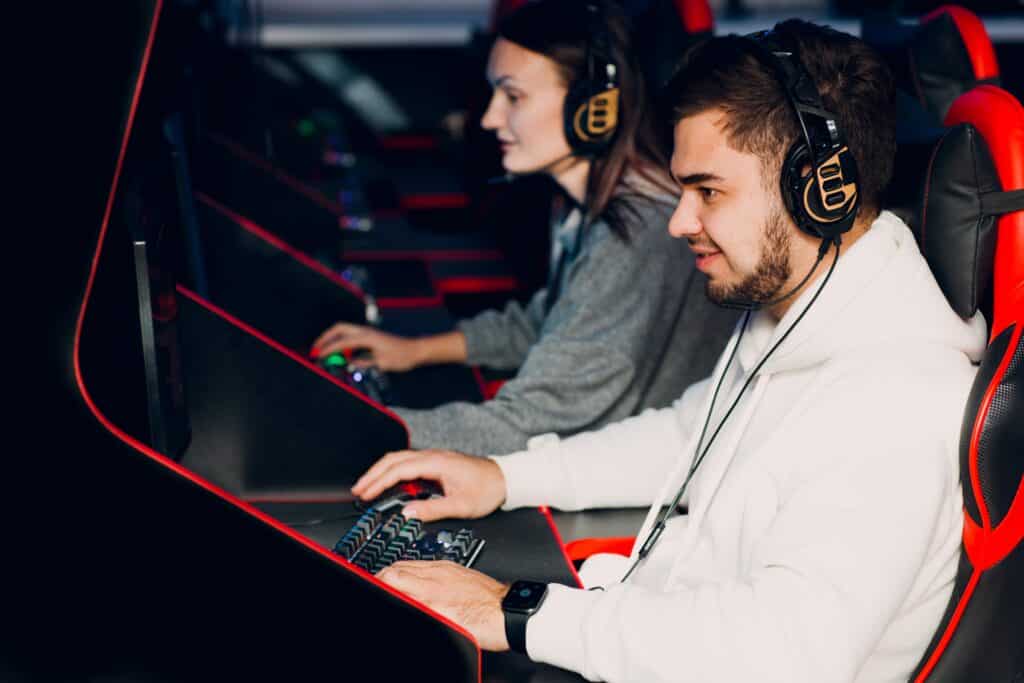 Um jovem de barba e moletom branco está sentado em uma cadeira gamer preta e vermelha, focado na tela de um computador. Ele usa fones de ouvido grandes e profissionais e opera um teclado mecânico iluminado. Ao lado dele, uma jovem também está equipada com fones de ouvido e joga em sua própria estação.