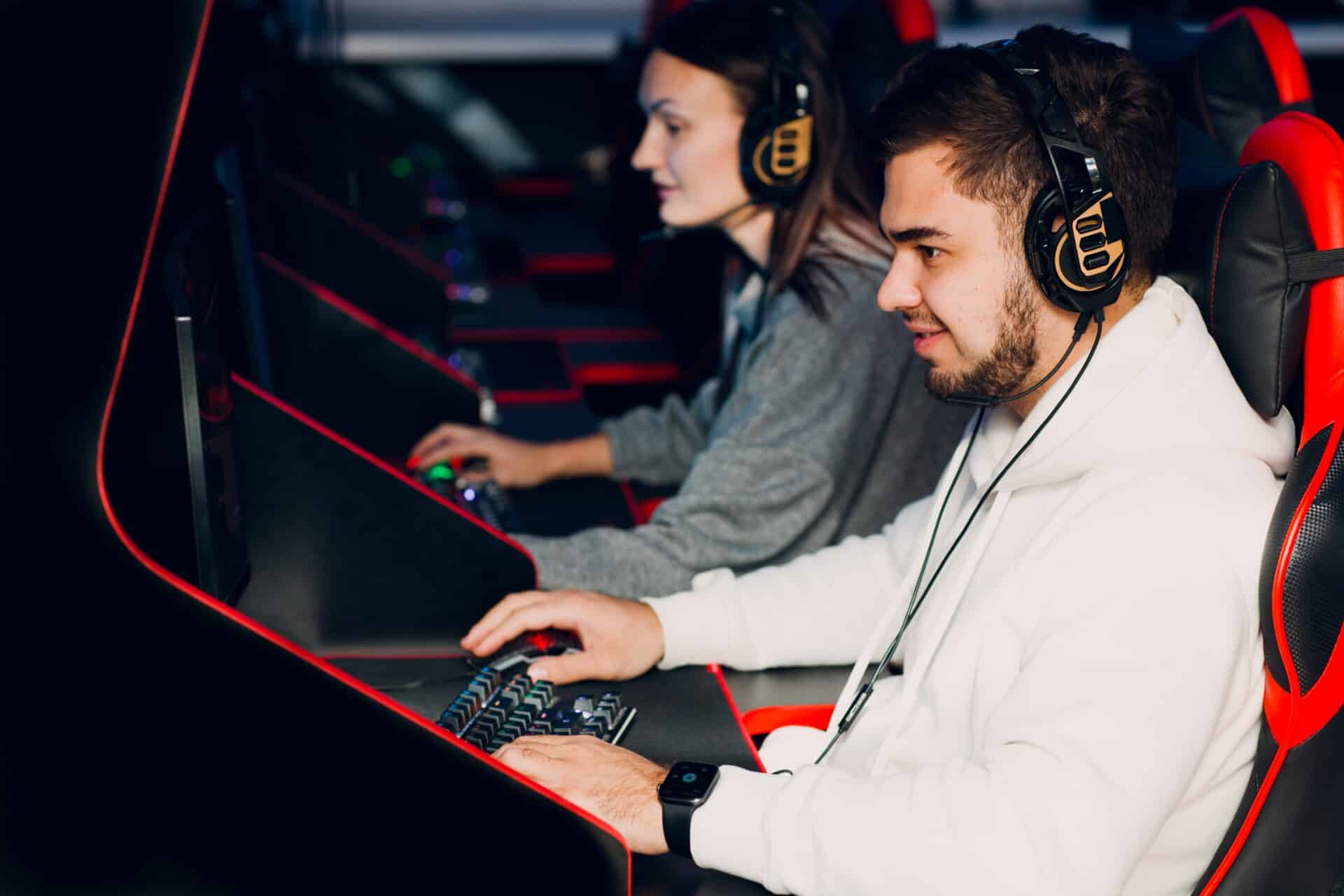 Um jovem de barba e moletom branco está sentado em uma cadeira gamer preta e vermelha, focado na tela de um computador. Ele usa fones de ouvido grandes e profissionais e opera um teclado mecânico iluminado. Ao lado dele, uma jovem também está equipada com fones de ouvido e joga em sua própria estação.