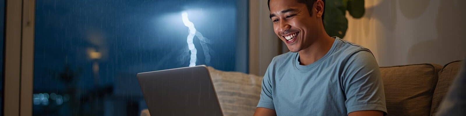 Um homem sorridente trabalha em um notebook em uma sala bem iluminada e confortável. Através de uma grande janela ao fundo, vê-se uma tempestade noturna com chuva forte e um raio iluminando o céu escuro