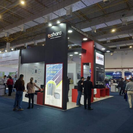Fotografia do estande da TS Shara em uma feira de negócios. O estande é moderno, com estrutura alta nas cores preto e vermelho e o logotipo iluminado no topo. Há grandes painéis verticais exibindo produtos, incluindo um destaque para a linha "TS Solar Off-Grid".