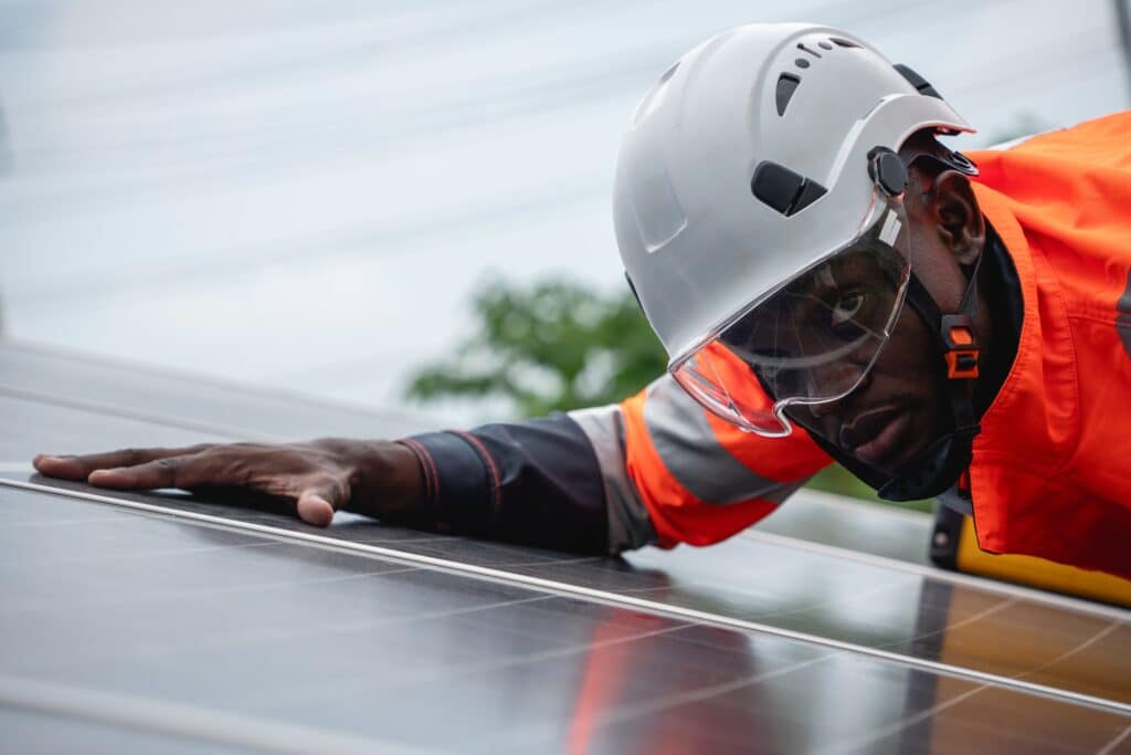 um engenheiro de serviço negro realizando a manutenção de painéis solares sob um céu nublado. Ele usa um capacete de segurança branco com viseira transparente e um uniforme laranja de alta visibilidade.