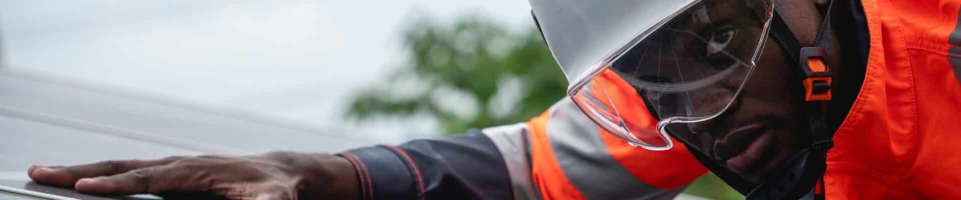 um engenheiro de serviço negro realizando a manutenção de painéis solares sob um céu nublado. Ele usa um capacete de segurança branco com viseira transparente e um uniforme laranja de alta visibilidade.