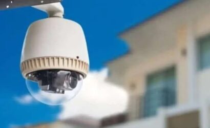 Câmera de segurança externa tipo dome instalada em uma parede, com fundo de céu azul e prédios desfocados.