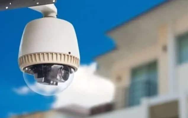 Câmera de segurança externa tipo dome instalada em uma parede, com fundo de céu azul e prédios desfocados.