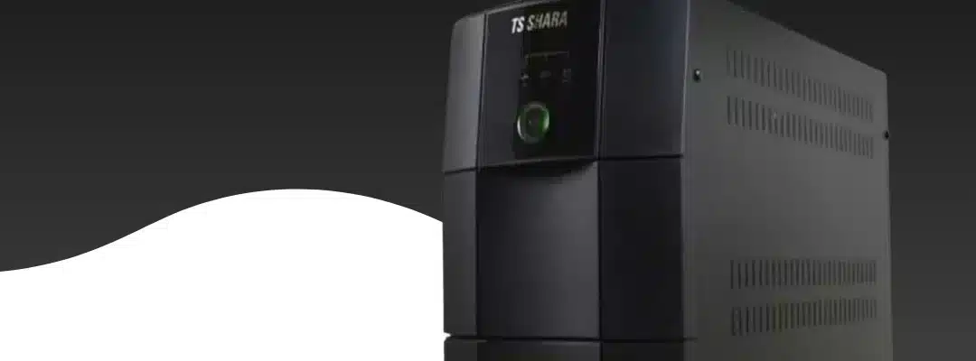 nobreak retangular preto da marca TS Shara, posicionado horizontalmente sobre uma superfície refletiva escura. O equipamento possui um design robusto com acabamento fosco e o logotipo da marca em destaque na parte frontal esquerda. À direita, há um painel com um botão de liga/desliga circular e indicadores de LED.