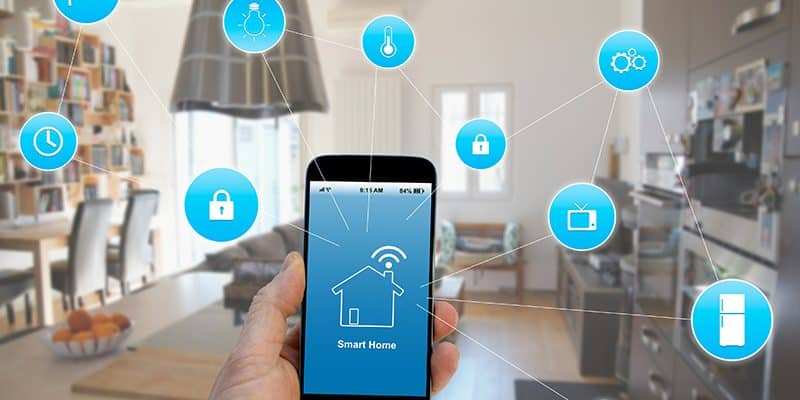 Uma mão segura um smartphone em primeiro plano, exibindo na tela um ícone de casa com sinal de Wi-Fi e a frase "Smart Home". Ao redor do celular, diversos ícones azuis flutuantes estão conectados por linhas finas, representando o controle remoto de dispositivos domésticos como câmeras de segurança, lâmpadas, termostatos, fechaduras, televisores e eletrodomésticos.