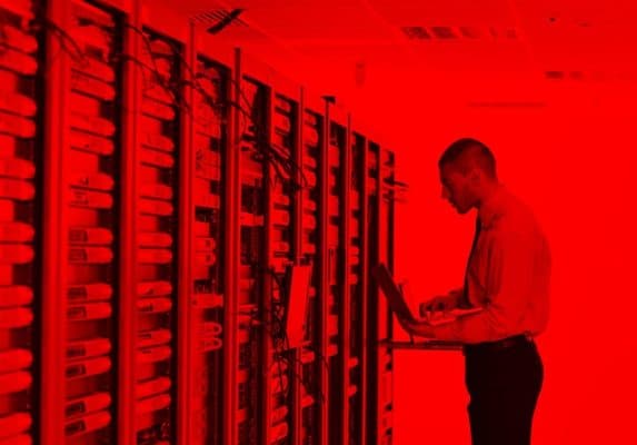 Engenheiro ou técnico de TI trabalhando com laptop em um Data Center ou sala de servidores, com o fundo com transparencia vermelha