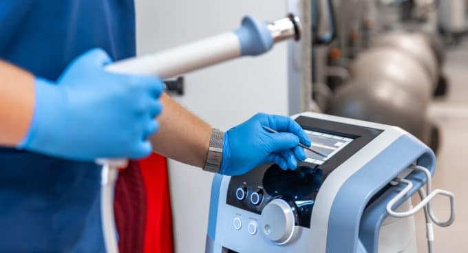 Profissional usando luvas azuis operando um equipamento médico ou fisioterapêutico com tela digital e um aplicador na mão