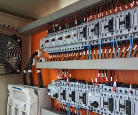 Detalhe de um painel elétrico ou quadro de distribuição industrial, com múltiplos disjuntores e fiação organizada, e um ventilador de resfriamento lateral