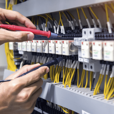 Mãos de técnico ou engenheiro realizando manutenção e teste de tensão em painel de controle industrial, utilizando pontas de prova em bornes e relés de proteção.