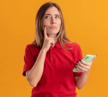Mulher jovem pensativa com camisa polo vermelha, segurando um smartphone verde e tocando o rosto com o dedo indicador, sobre fundo laranja.