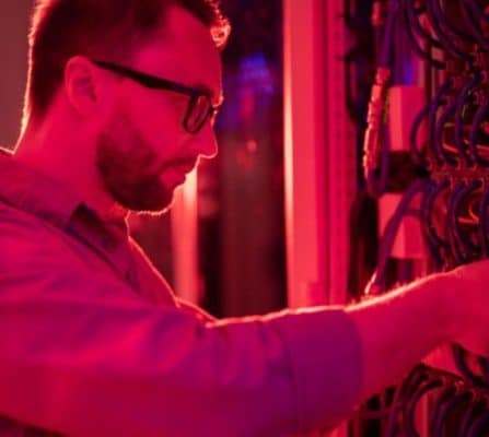 Homem com barba e óculos trabalhando em um rack de servidor, em ambiente de data center com iluminação vermelha.