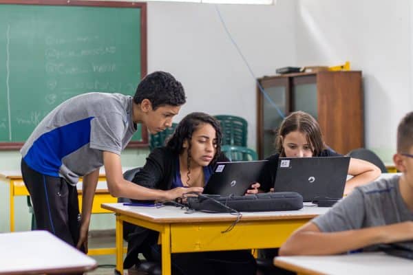 Três estudantes adolescentes utilizando laptops em sala de aula com quadros-negros ao fundo