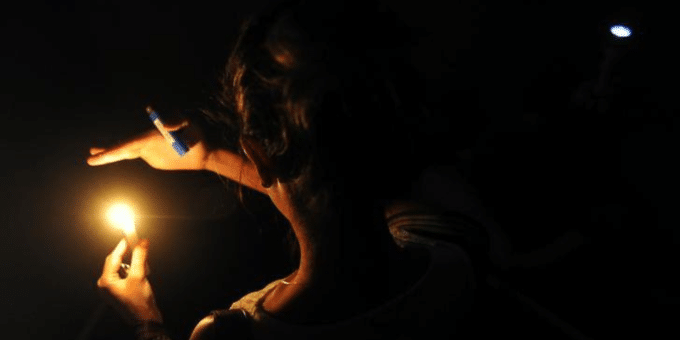 Uma mulher em um ambiente totalmente escuro. A pessoa está usando uma regata clara e ilumina o ambiente com uma única fonte de luz em sua mão: uma vela ou um isqueiro aceso