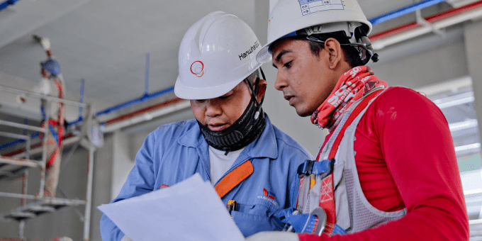 Dois homens, usando capacetes de segurança brancos e coletes de segurança, estão lado a lado e concentrados em analisar um documento branco que seguram nas mãos