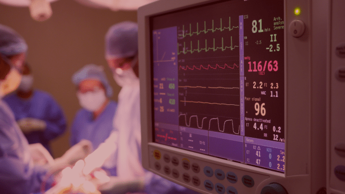 Monitor cardíaco exibindo sinais vitais de um paciente durante uma cirurgia, com equipe médica usando máscaras e roupas estéreis ao fundo
