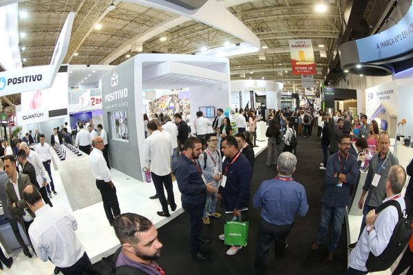 Vista ampla e superior de um evento ou feira de tecnologia, mostrando um pavilhão movimentado com grande público.