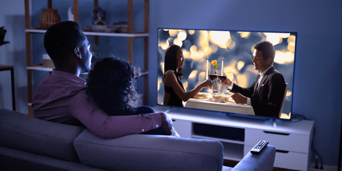 Um casal, visto por trás, sentados em um sofá em uma sala escura, assistindo televisão juntos, o que parece ser um filme de romance 