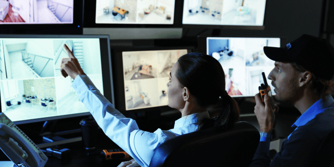A imagem mostra dois profissionais de segurança em uma sala de monitoramento observando várias telas com imagens de câmeras de vigilância. Uma mulher aponta para um dos monitores enquanto um homem ao lado segura um rádio comunicador.
