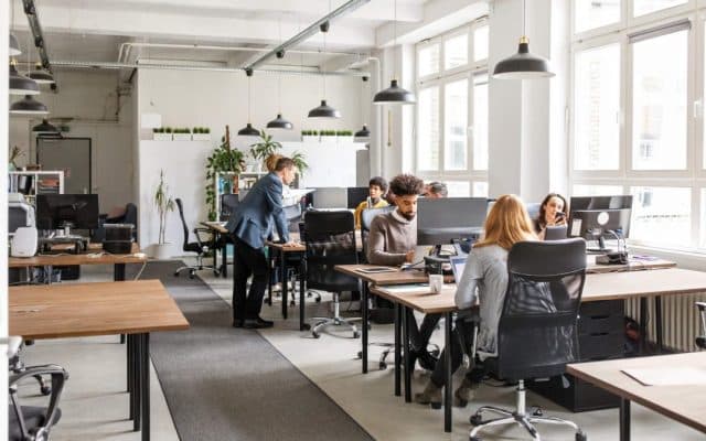 escritório moderno com várias pessoas trabalhando em mesas compartilhadas. O ambiente é iluminado por luz natural vinda de grandes janelas e por luminárias pendentes no teto. Há computadores sobre as mesas, cadeiras ergonômicas e plantas decorativas espalhadas pelo espaço.