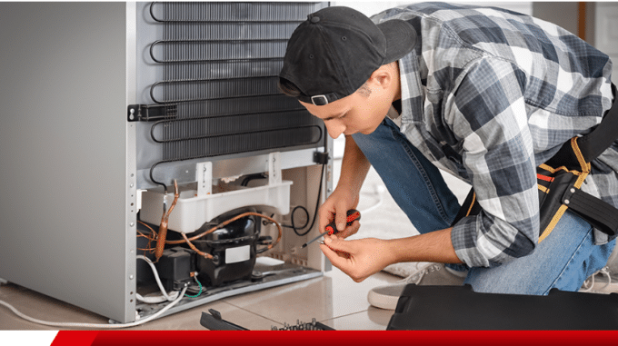 Um jovem técnico, usando um boné preto virado para trás, camisa xadrez e calça jeans, está agachado consertando a parte traseira de uma geladeira