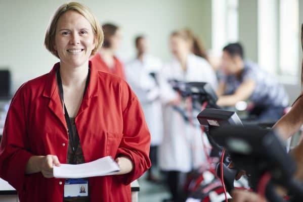 Mulher sorridente de jaleco vermelho segura papéis em uma sala com pessoas ao fundo