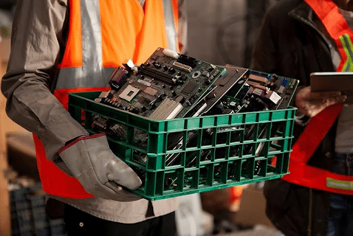 Uma pessoa vestindo luvas de trabalho e um colete de segurança laranja sobre uma camisa cinza segura uma caixa plástica verde cheia de placas de circuito eletrônicas
