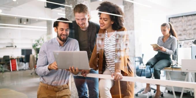 Três jovens profissionais (dois homens e uma mulher) em um escritório, estão reunidos e sorrindo enquanto olham para a tela de um laptop que um deles segura