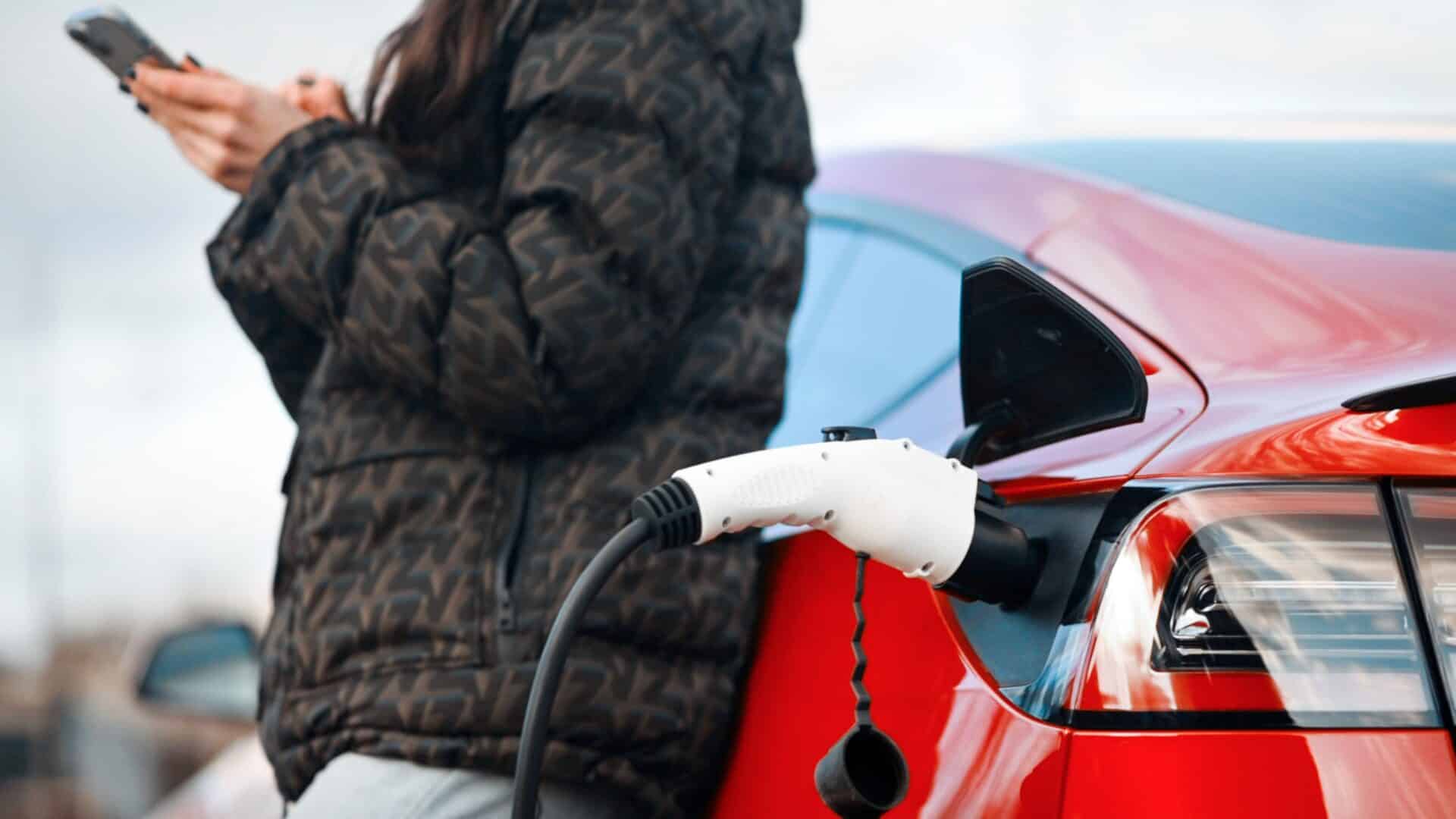 Uma pessoa em pé ao lado de um carro elétrico vermelho que está sendo carregado. O foco da imagem está no bocal de carregamento branco conectado à lateral traseira do veículo.