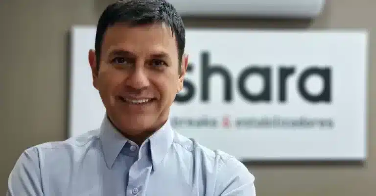 Homem sorridente de camisa social clara, em frente a um letreiro com o nome “Shara” parcialmente visível.