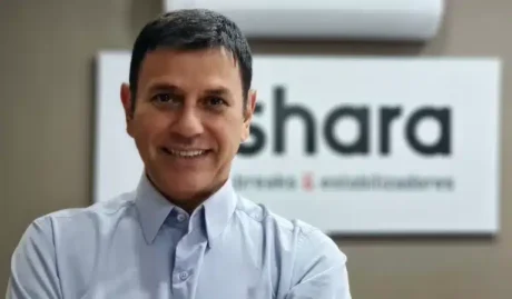 Homem sorridente de camisa social clara, em frente a um letreiro com o nome “Shara” parcialmente visível.