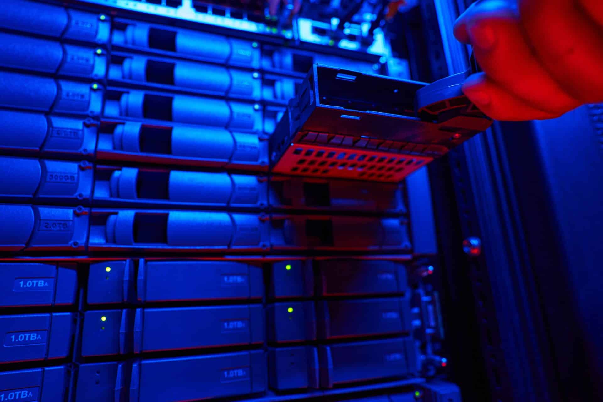 um rack de servidores repleto de discos rígidos (HDDs) organizados em gavetas. A imagem é iluminada por uma forte luz azul neon, com detalhes em vermelho. Uma mão humana aparece no canto superior direito, deslizando uma das gavetas para fora do compartimento.