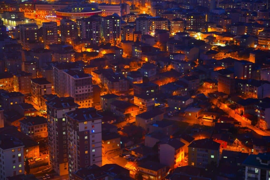 A imagem é uma vista aérea noturna de uma grande área urbana. Milhares de luzes alaranjadas, vindas das janelas dos edifícios e das lâmpadas das ruas, iluminam a paisagem e criam um forte contraste com o céu escuro. As construções variam em altura, com alguns prédios mais altos e muitos outros de menor porte, formando um visual denso e intrincado que se estende por toda a foto.