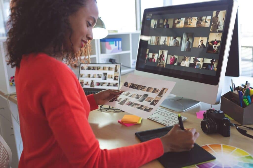 Uma mulher com cabelos encaracolados escuros, vestindo uma blusa vermelha, está sentada em uma mesa de escritório, trabalhando em um computador. Em sua mesa, há dois monitores, um deles grande e exibindo uma grade de fotos. A mulher segura uma folha com várias miniaturas de fotos e, com a mão direita, usa uma caneta stylus em uma mesa digitalizadora. Vários objetos de escritório, como um mouse, um teclado, uma câmera e uma roda de cores, também estão na mesa, sugerindo que ela trabalha com design gráfico ou edição de fotos.