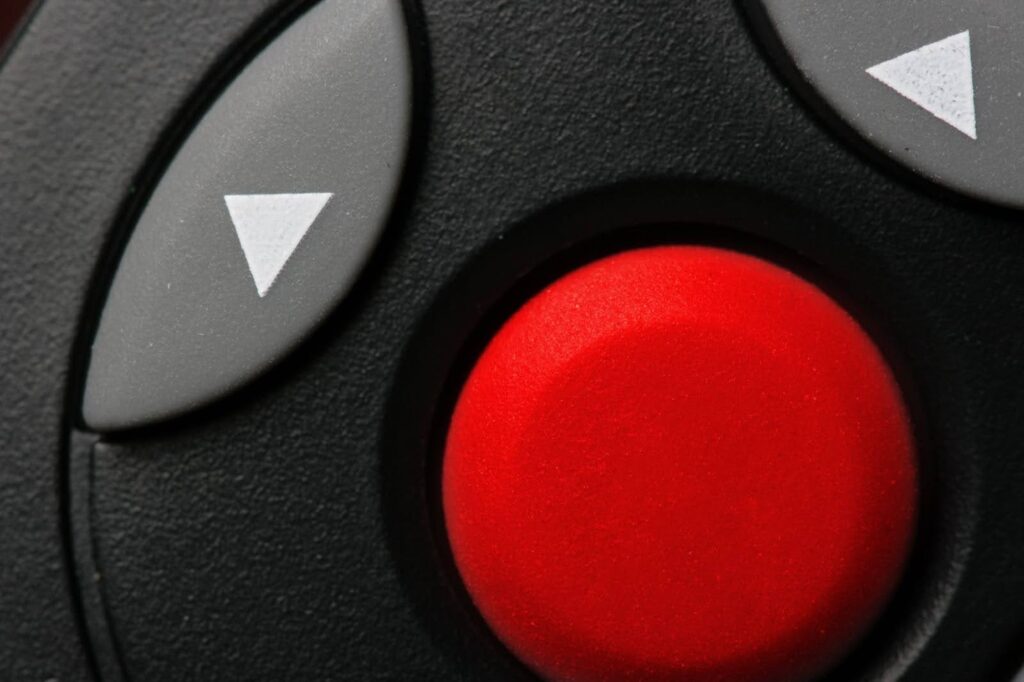 A segunda imagem é um close-up extremo do centro de um controle remoto ou joystick de cor preta. O ponto focal é um grande botão circular de cor vermelha, que se destaca em alto-relevo no meio da imagem. Ao seu redor, há botões em forma de flechas brancas e cinza escuro. As flechas apontam para cima e para baixo no lado esquerdo e para a direita no lado superior direito. O foco na imagem está no botão vermelho, que parece ser o principal ponto de controle.