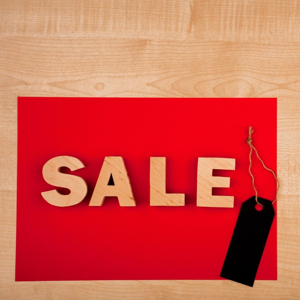 Sobre uma superfície de madeira clara, há um retângulo vermelho colocado como fundo. Em cima dele, a palavra “SALE” (em inglês, “promoção” ou “liquidação”) está escrita em letras grandes de madeira, posicionadas lado a lado. À direita, preso por um cordão, há um marcador preto em formato de etiqueta de preço, reforçando a ideia de desconto ou oferta.