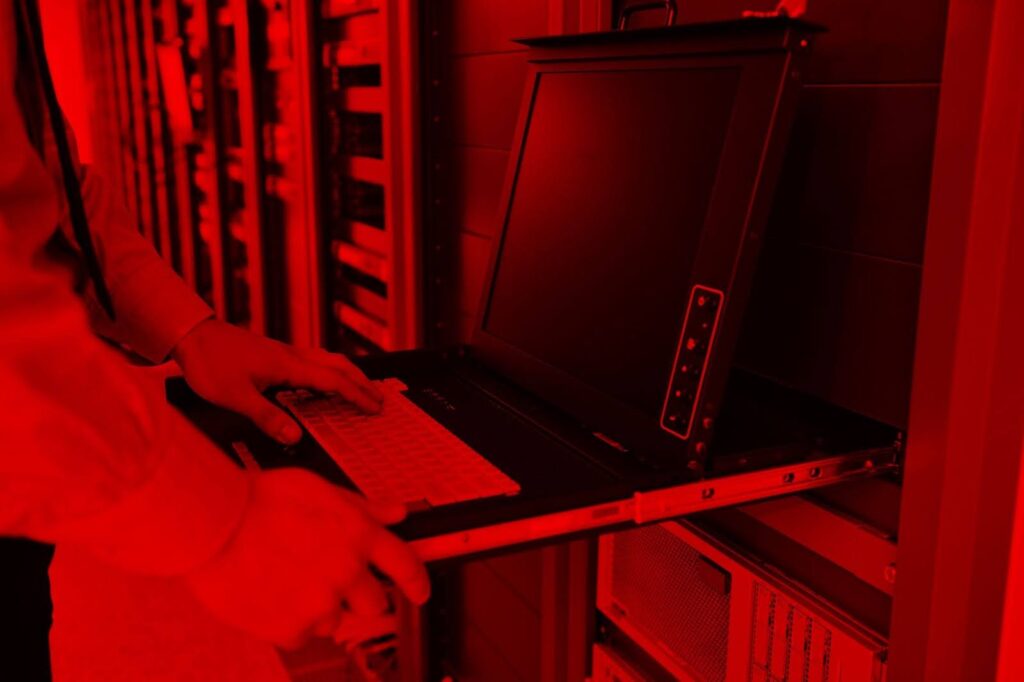 Um ambiente de servidores de tecnologia. Uma pessoa, visível apenas das mãos e parte do corpo, está puxando um console retrátil com teclado e monitor embutidos, usado para acessar servidores. Toda a cena está coberta por um filtro vermelho intenso, que cria um clima tecnológico e moderno.