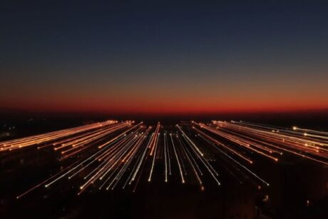 Um horizonte ao entardecer, com o céu em tons de azul escuro na parte superior e alaranjado próximo ao horizonte. Da paisagem urbana, partem múltiplos feixes de luz em linhas retas, que se estendem para frente como trilhas luminosas. O efeito cria a sensação de movimento acelerado ou de visão futurista da cidade.