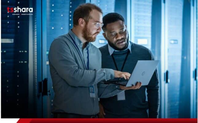 Dois profissionais de TI analisando dados em notebook dentro de data center.