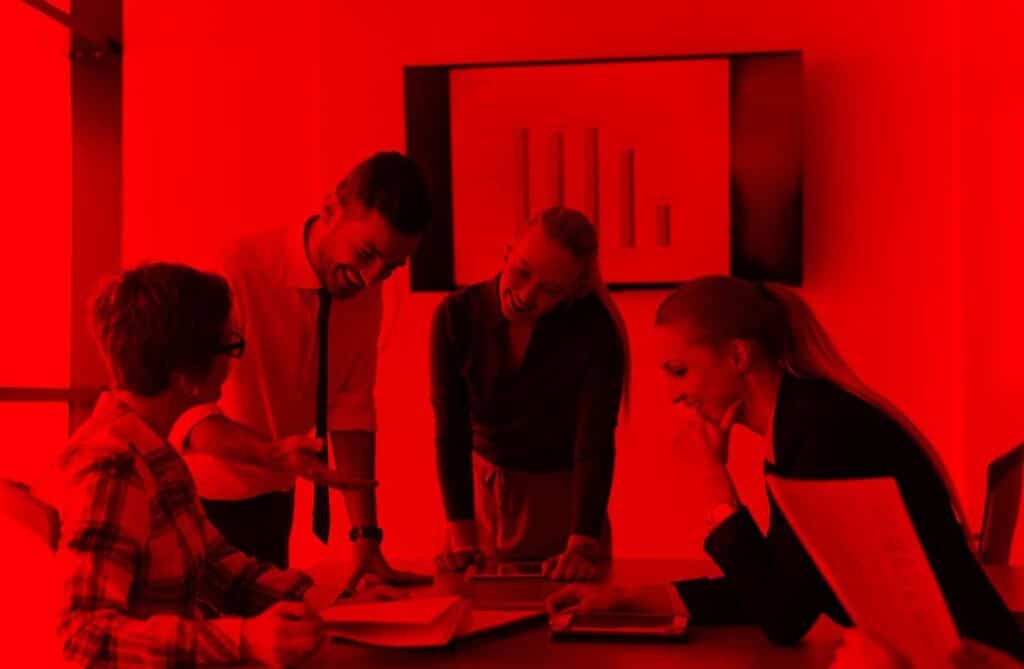 Quatro pessoas estão reunidas em uma sala de reunião, ao redor de uma mesa. Duas mulheres e dois homens, todos em roupas formais, conversam animadamente enquanto olham para documentos e um notebook sobre a mesa. Ao fundo, uma tela exibe um gráfico de barras. Toda a imagem está coberta por um filtro vermelho intenso, transmitindo uma atmosfera de energia e intensidade.