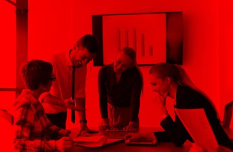 Quatro pessoas estão reunidas em uma sala de reunião, ao redor de uma mesa. Duas mulheres e dois homens, todos em roupas formais, conversam animadamente enquanto olham para documentos e um notebook sobre a mesa. Ao fundo, uma tela exibe um gráfico de barras. Toda a imagem está coberta por um filtro vermelho intenso, transmitindo uma atmosfera de energia e intensidade.